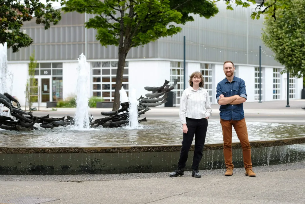 Sterner Stuff owner Ethan Clevenger and associate developer Maddy Harrison pose in front of the water feature in the Park Blocks in Downtown Eugene, Oregon. The Farmers Market Pavilion can be seen in the background.